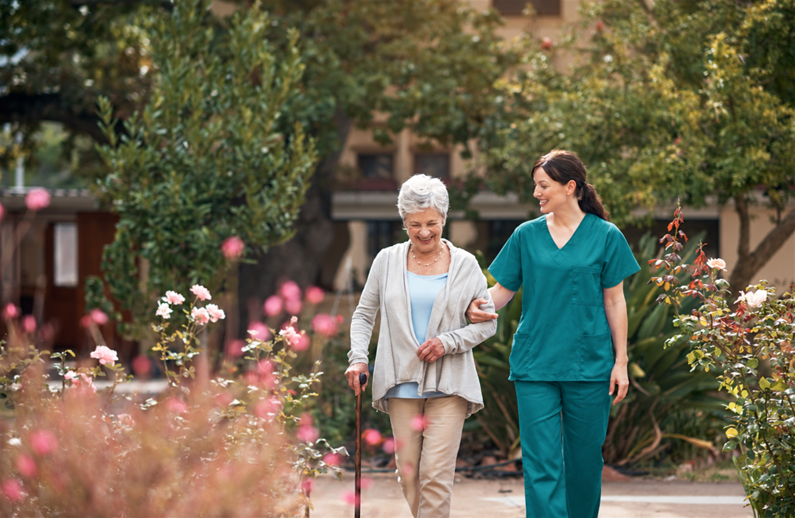A care worker and older woman walking together through a garden.
